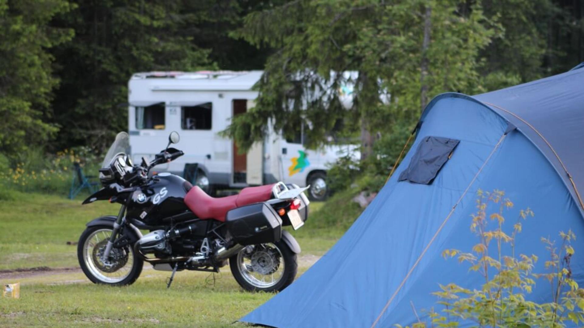 Campeggio sereno con moto, tenda blu e camper tra alberi verdi.